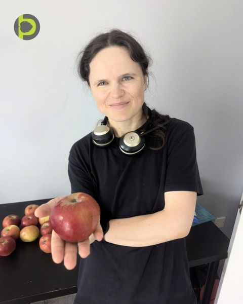Dr Maryna Kuzmenko in a black t-shirt holding out a red apple, with more apples on a dark table behind her.