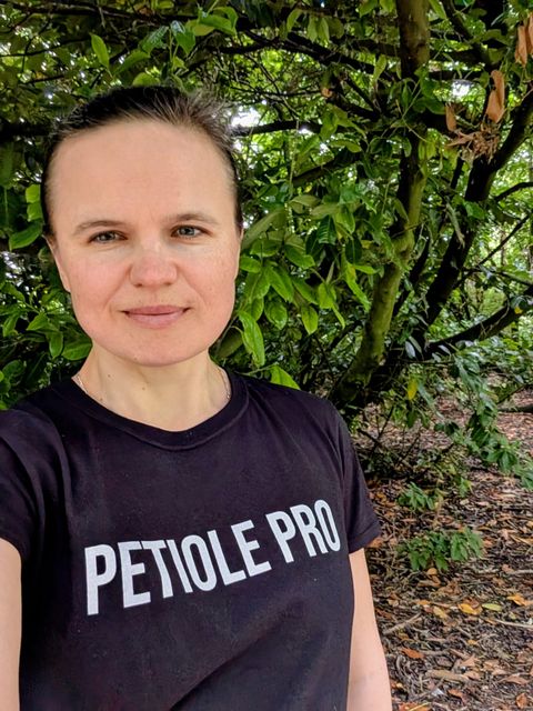 Dr Maryna Kuzmenko, founder of Petiole Pro, standing in front of dense green foliage wearing a black Petiole Pro t-shirt.