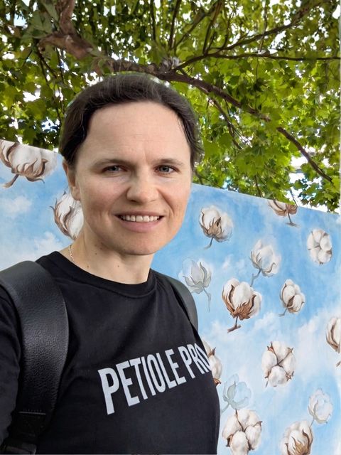 Dr Maryna Kuzmenko wearing a black Petiole Pro t-shirt, standing under a tree in front of a painted backdrop of cotton bolls against a blue sky.